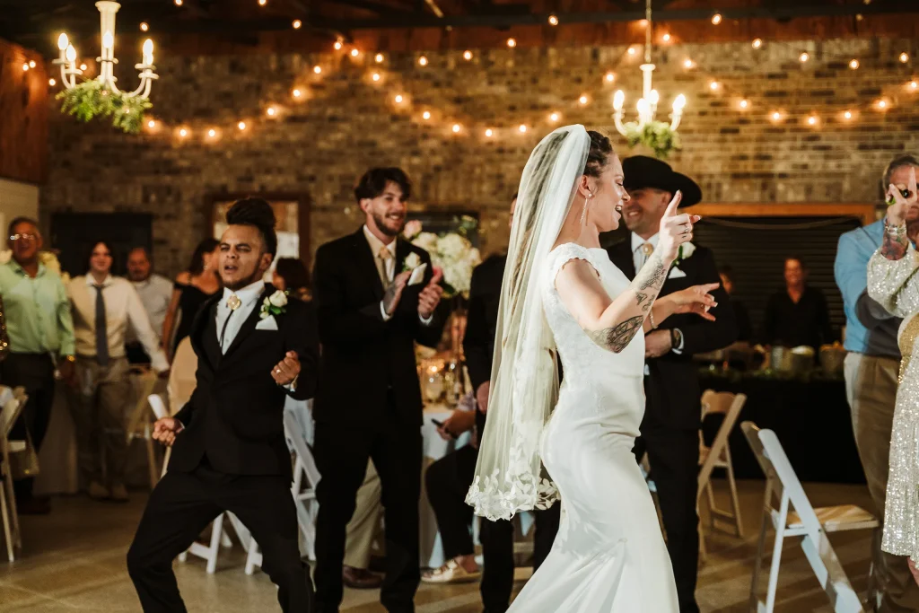 The bride and groom dance joyfully during their wedding reception at Masters Stables, surrounded by guests and warm string lights.