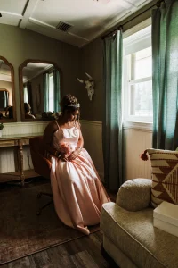 Bridesmaid in a pink gown sitting by the window in the Masters Stables bridal suite before the ceremony.