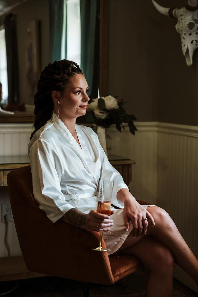 Bride sitting in the Masters Stables bridal suite with a champagne glass, wearing a white robe while preparing for her wedding day in Fellsmere, Florida.