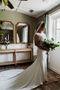 The bride stands in the Masters Stables bridal suite holding her bouquet, illuminated by natural window light with her train flowing behind her.