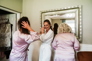 Bride and her bridesmaids getting ready in the Masters Stables bridal suite, adjusting hair and earrings in front of a large mirror.