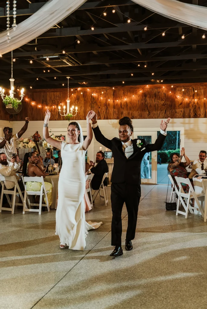 The bride and groom make their grand entrance into the reception hall at Masters Stables, smiling and raising their hands as guests cheer.