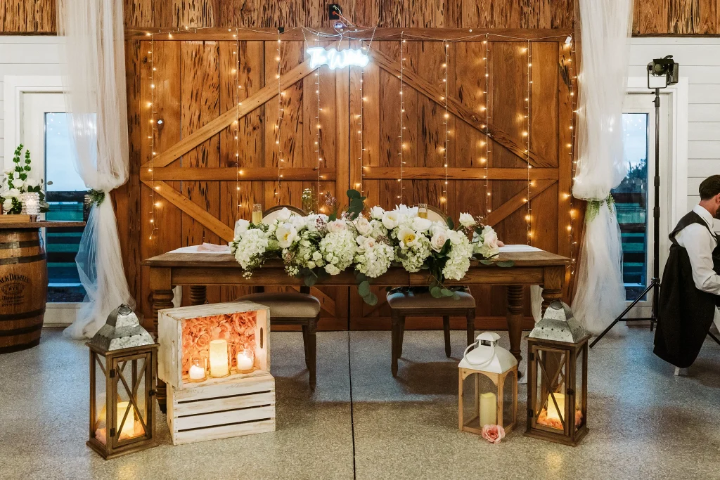 Elegant sweetheart table decorated with white and blush florals, lanterns, and string lights inside the Masters Stables wedding reception barn.
