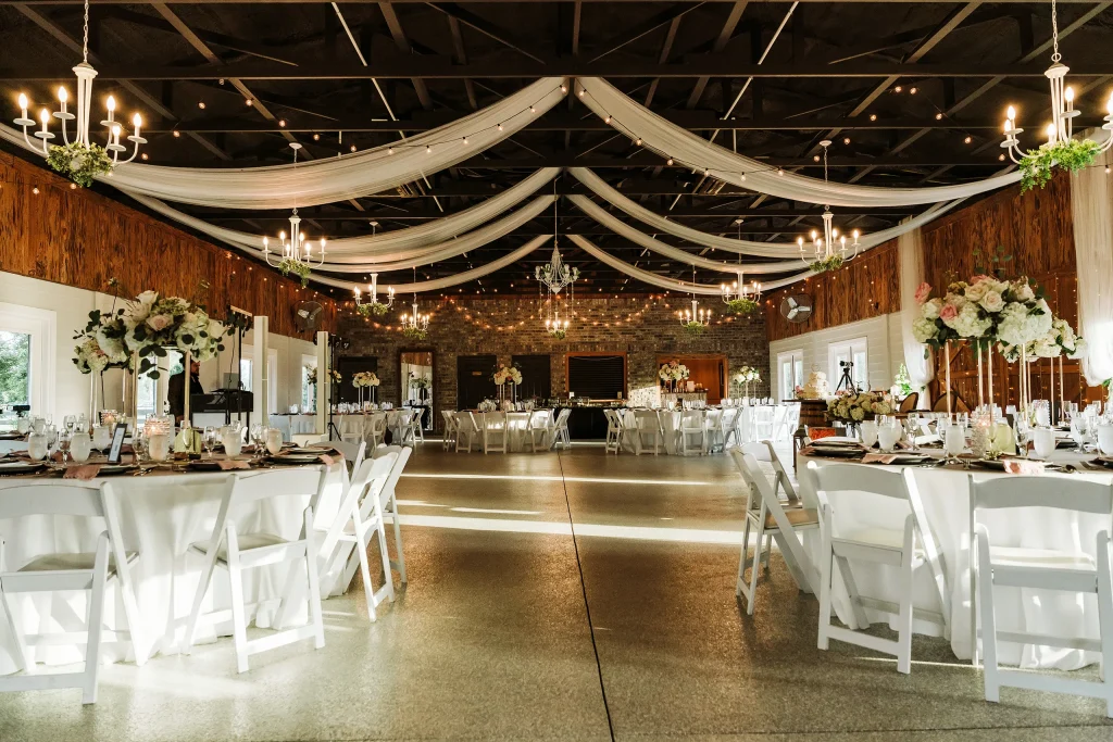 Wide view of the decorated wedding reception hall at Masters Stables in Fellsmere, Florida, featuring draped ceilings, chandeliers, round tables, and elegant floral centerpieces.