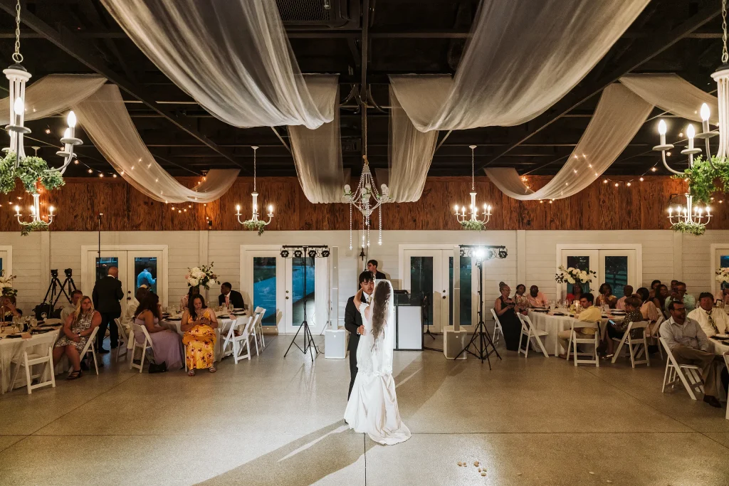 Bride and groom sharing their first dance inside the beautifully decorated reception hall at Masters Stables in Fellsmere, Florida, with draped ceilings, chandeliers, and wedding guests seated around them.