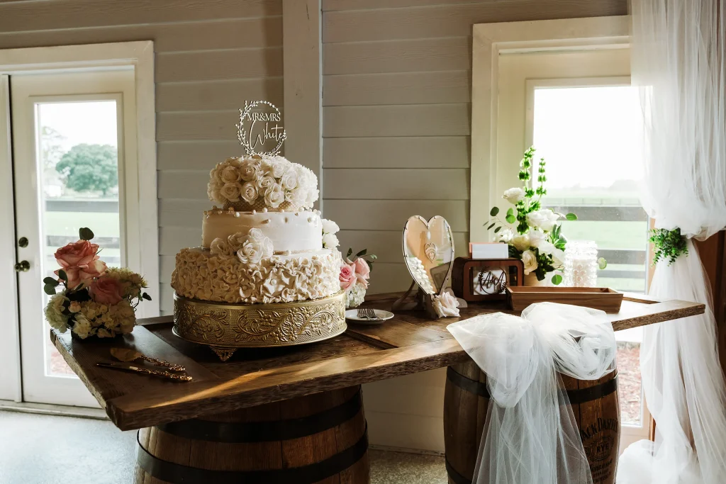 Elegant wedding cake table displayed on whiskey barrels at Masters Stables, featuring a tiered white cake, floral d&eacute;cor, and a heart-shaped guest sign.