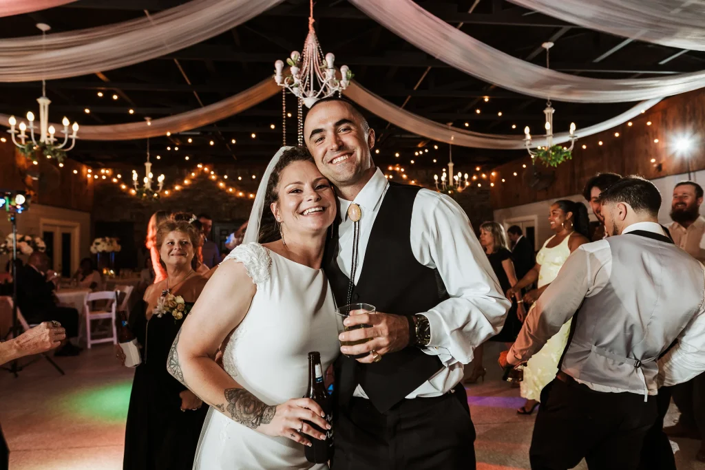 Bride smiling and posing with a groomsman during the wedding reception at Masters Stables, surrounded by guests and twinkle-light d&eacute;cor.